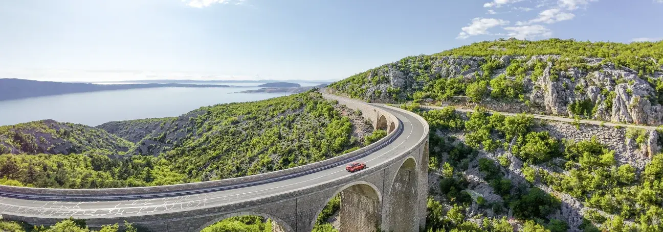[Drohnenfoto eines roten Fahrzeugs auf Viadrukt durch mediterane Landschaft]