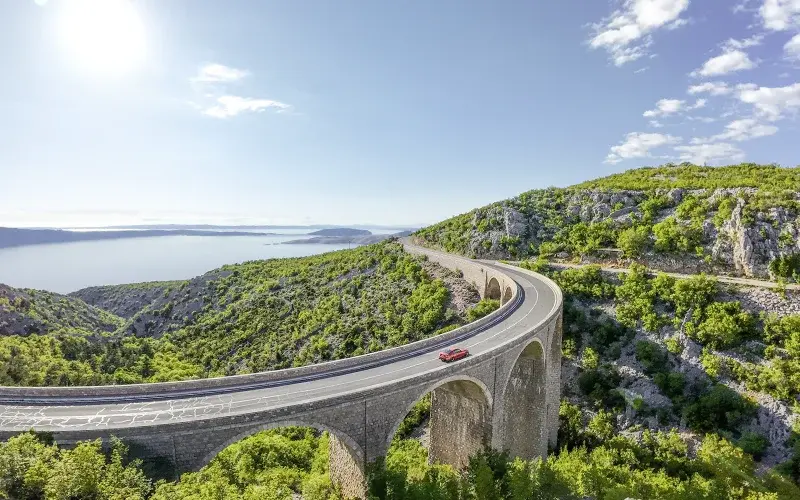 [Drohnenfoto eines roten Fahrzeugs auf Viadrukt durch mediterane Landschaft]